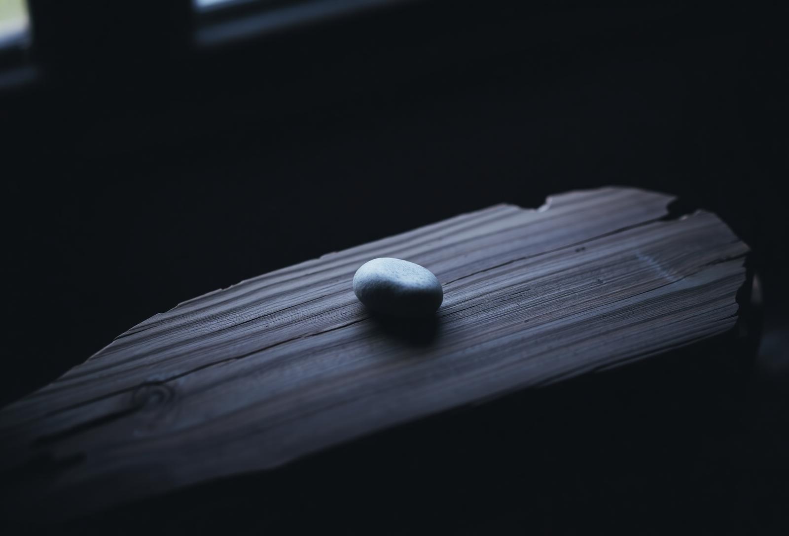 A single stone resting on weathered wood in soft window light, evoking patience and depth.